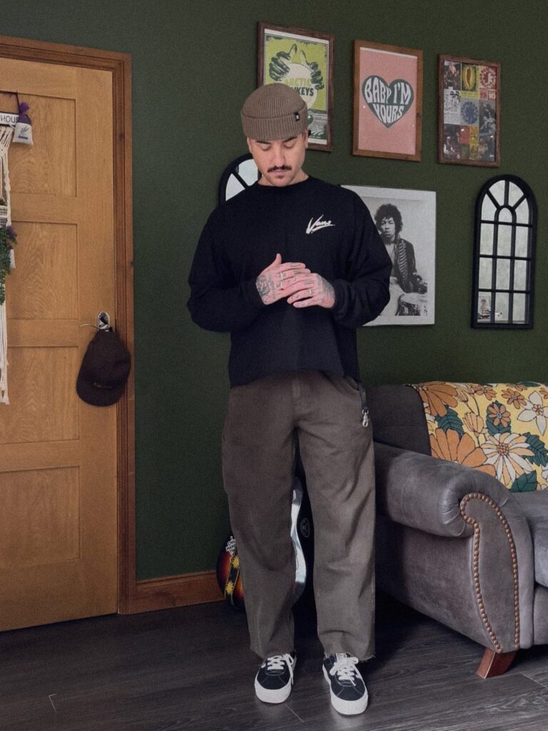 A man wearing baggy brown pants, a black shirt, and a cap stands in a room.