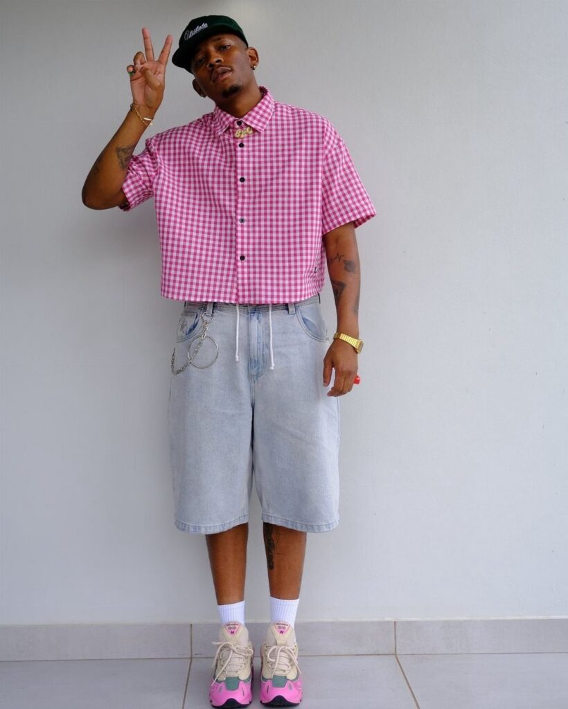 Man in pink checkered shirt and denim shorts posing against a white wall.