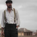 A bearded man wearing a flat cap, striped shirt, and suspenders standing outdoors.