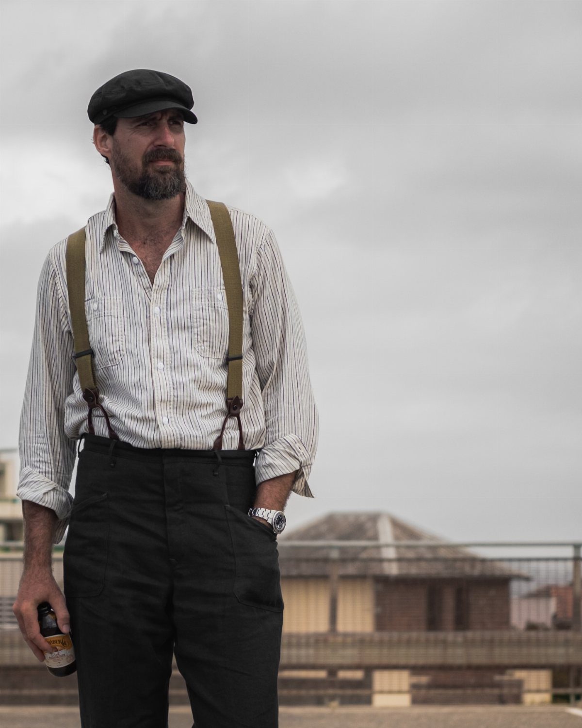 A bearded man wearing a flat cap, striped shirt, and suspenders standing outdoors.